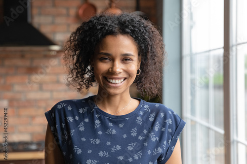 Portrait of happy millennial Black mixed race girl standing by window at home, looking at camera with toothy smile. Beautiful African American young woman, female model in house interior. Head shot
