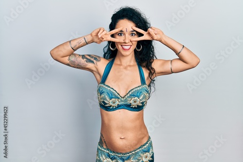 Young woman wearing bindi and traditional belly dance clothes doing peace symbol with fingers over face, smiling cheerful showing victory © Krakenimages.com