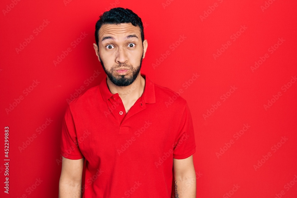 Hispanic man with beard wearing casual red t shirt puffing cheeks with funny face. mouth inflated with air, crazy expression. Hispanic man with beard wearing casual red t shirt puffing cheeks with funny face. mouth inflated with air, crazy expression.