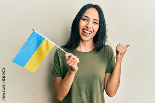 Young hispanic girl wearing ukraine flag pointing thumb up to the side smiling happy with open mouth
