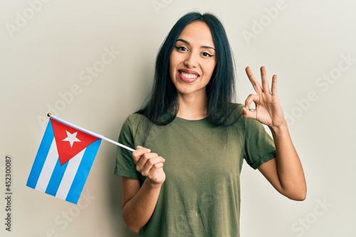 Young hispanic girl holding cuba flag doing ok sign with fingers, smiling friendly gesturing excellent symbol