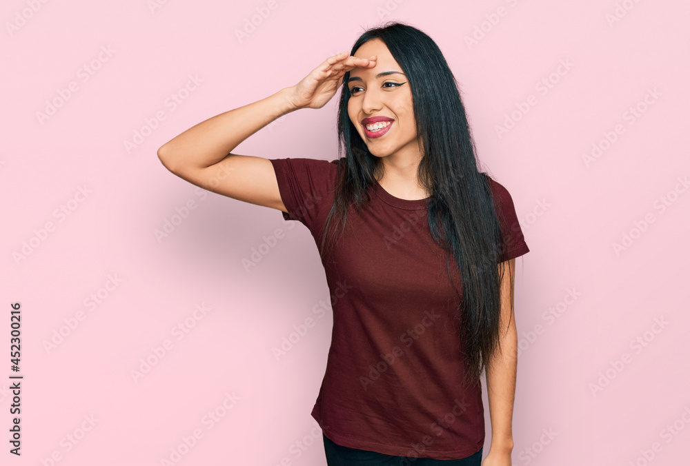 Young hispanic girl wearing casual t shirt very happy and smiling looking far away with hand over head. searching concept. Young hispanic girl wearing casual t shirt very happy and smiling looking far away with hand over head. searching concept.