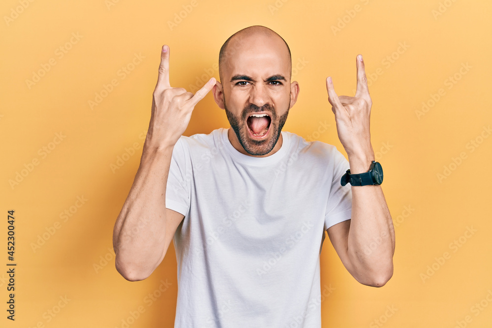 Fototapeta premium Young bald man wearing casual white t shirt shouting with crazy expression doing rock symbol with hands up. music star. heavy music concept.