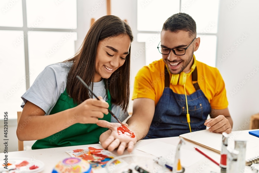 Fototapeta premium Young hispanic couple smiling happy painting hands sitting on the table at art studio.