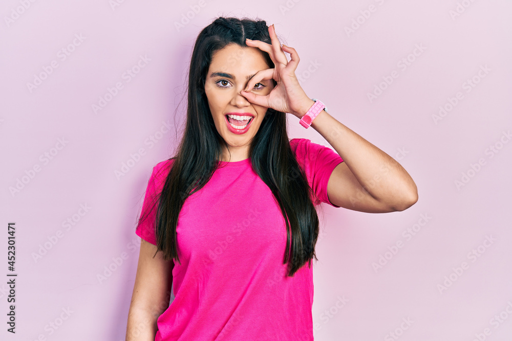 Young hispanic girl wearing casual pink t shirt doing ok gesture with hand smiling, eye looking through fingers with happy face. Young hispanic girl wearing casual pink t shirt doing ok gesture with hand smiling, eye looking through fingers with happy face.