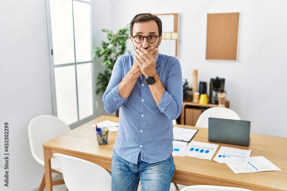 Middle age hispanic man with beard wearing business clothes at the office shocked covering mouth with hands for mistake. secret concept. Middle age hispanic man with beard wearing business clothes at the office shocked covering mouth with hands for mistake. secret concept.