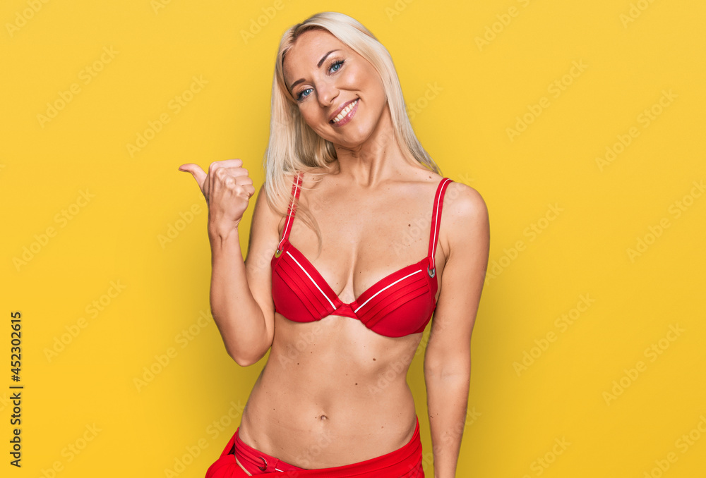 Naklejka premium Young blonde woman wearing bikini smiling with happy face looking and pointing to the side with thumb up.