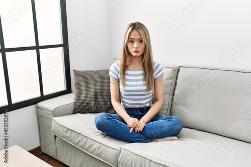 Asian young woman sitting on the sofa at home relaxed with serious expression on face. simple and natural looking at the camera.