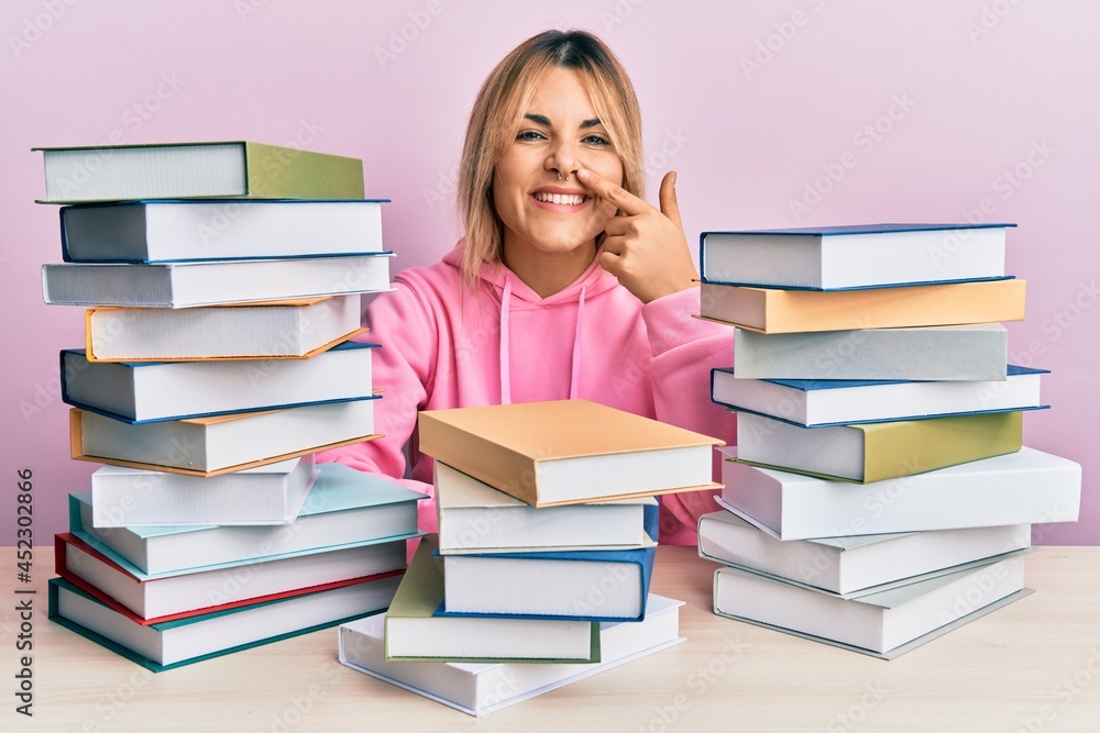 Young caucasian woman sitting on the table with books pointing with hand finger to face and nose, smiling cheerful. beauty concept Young caucasian woman sitting on the table with books pointing with hand finger to face and nose, smiling cheerful. beauty concept