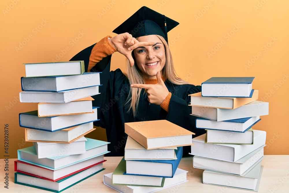 Young caucasian woman wearing graduation ceremony robe sitting on the table smiling making frame with hands and fingers with happy face. creativity and photography concept. Young caucasian woman wearing graduation ceremony robe sitting on the table smiling making frame with hands and fingers with happy face. creativity and photography concept.