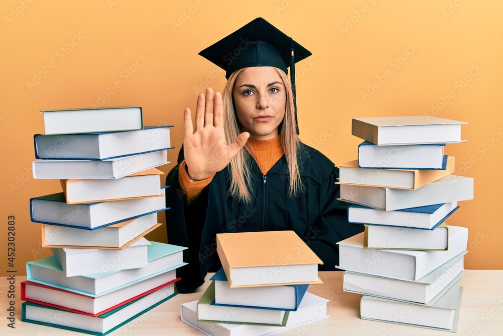 Young caucasian woman wearing graduation ceremony robe sitting on the table doing stop sing with palm of the hand. warning expression with negative and serious gesture on the face.