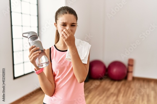 Young brunette teenager wearing sportswear holding water bottle smelling something stinky and disgusting, intolerable smell, holding breath with fingers on nose. bad smell