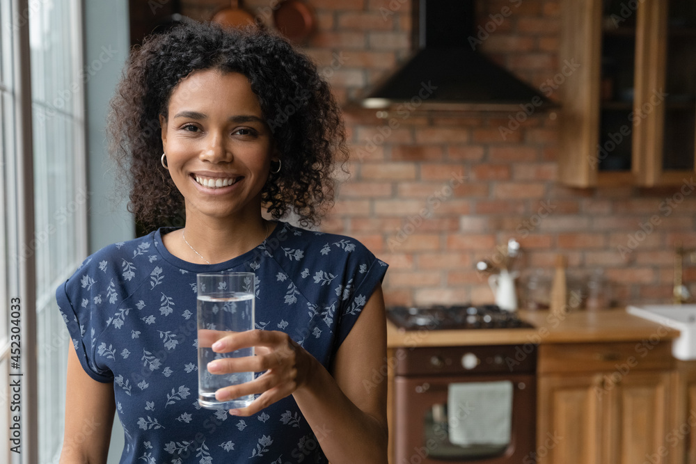 Happy young African mixed race woman drinking fresh pure natural water in kitchen, holding transparent glass, looking, smiling at camera with toothy smile, keeping healthy habit. Head shot portrait Happy young African mixed race woman drinking fresh pure natural water in kitchen, holding transparent glass, looking, smiling at camera with toothy smile, keeping healthy habit. Head shot portrait