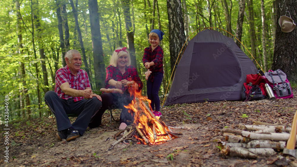 Senior elderly grandmother grandfather with granddaughter cooking frying sausages over campfire in wood. Old man woman and child girl kid tourists having camping in forest. Active rest of family Senior elderly grandmother grandfather with granddaughter cooking frying sausages over campfire in wood. Old man woman and child girl kid tourists having camping in forest. Active rest of family