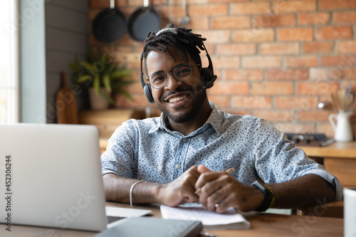 Portrait of happy mixed race male student in headset satisfied with distance education, virtual training, online webinar. College guy studying from home, writing notes, smiling at camera, Head shot