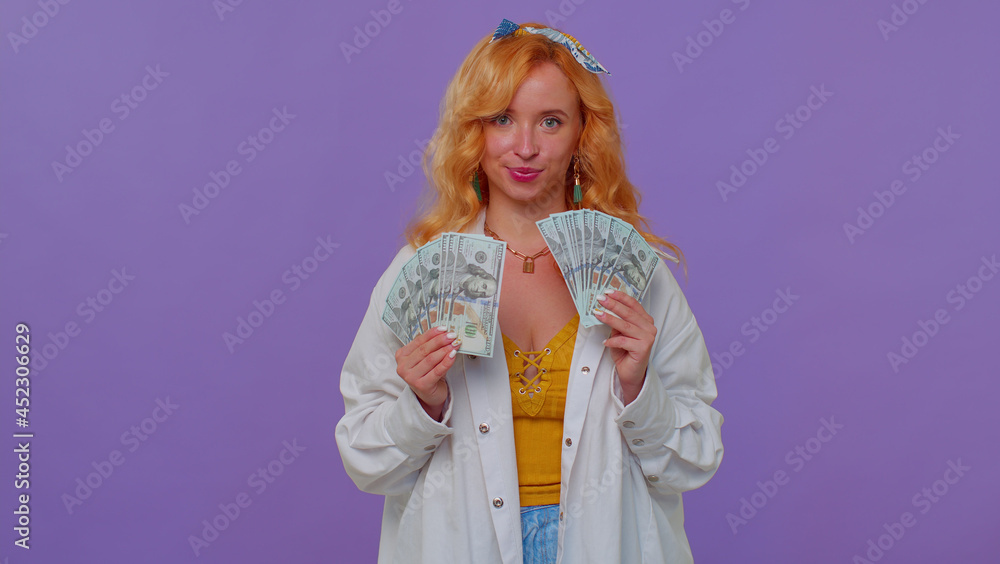 Arrogant rich stylish girl in white shirt holding money cash in dollars banknotes, smiling and looking at camera. Isolated over purple studio wall background. Concept of income, credit, debt, savings Arrogant rich stylish girl in white shirt holding money cash in dollars banknotes, smiling and looking at camera. Isolated over purple studio wall background. Concept of income, credit, debt, savings