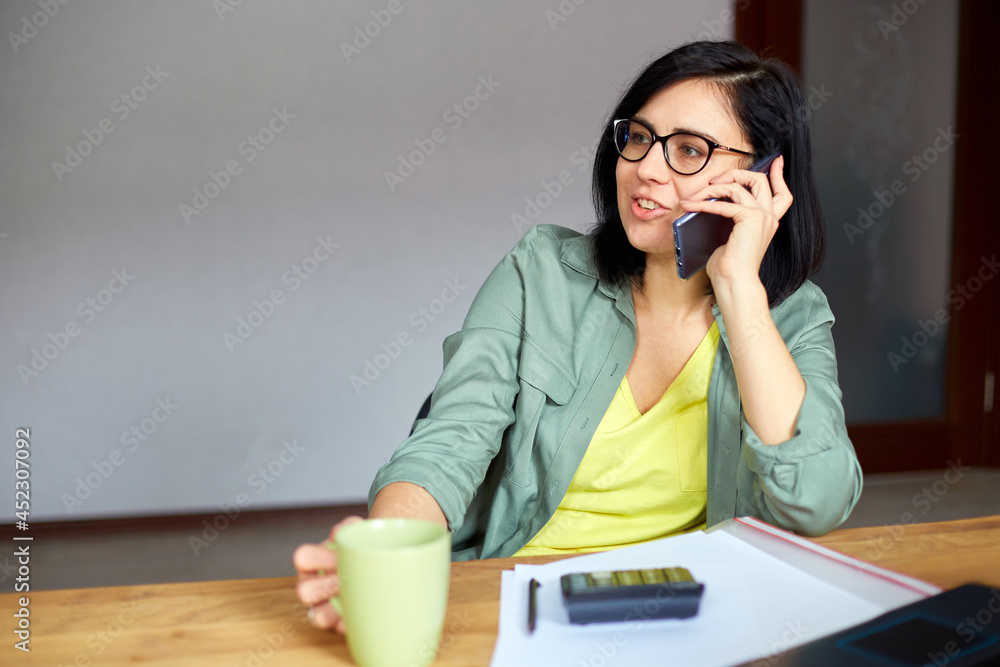 Stylish brunette woman in glasses sitting at wooden table with notepad and having phone call Stylish brunette woman in glasses sitting at wooden table with notepad and having phone call