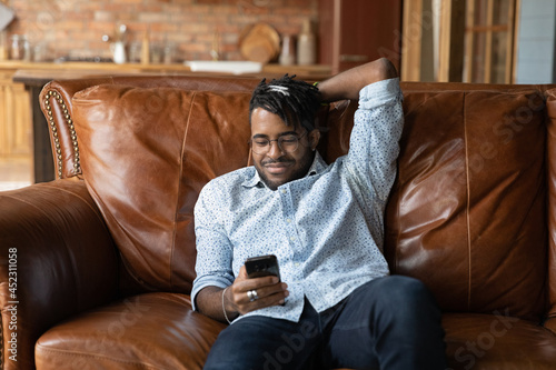 Cuadro en lienzo Happy lazy African American guy using smartphone, enjoying leisure at home, relaxing on leather couch, overusing mobile phone, texting message, chatting online