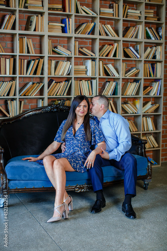 stylish man in a blue shirt with a beautiful wife in a dress are sitting together on a cozy sofa against the background of a luxurious room with books. Family happiness