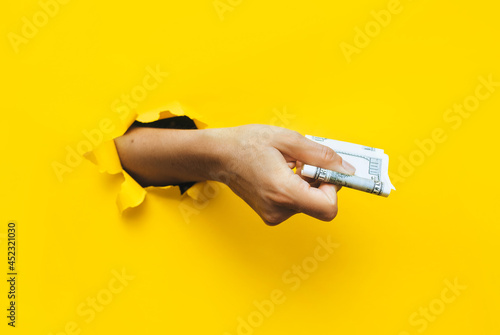A female hand appears in the hole in torn yellow paper and squeezes two hundred-dollar bills. The concept of poverty, benefits, scholarships, and stinginess. Isolated. Copy space. photo
