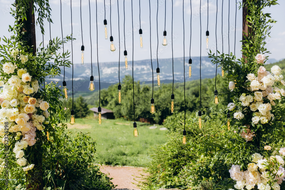 Fototapeta premium Wedding arch on rustuc style with white flowers in park with light bulbs.