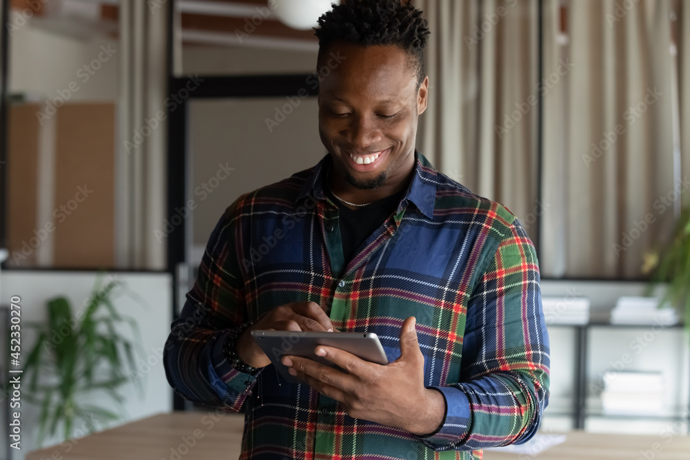 Obraz premium Smiling young African American businessman use tablet text message online on gadget. Happy millennial mixed race male employee hold pad device communicate with client on internet. Technology concept.