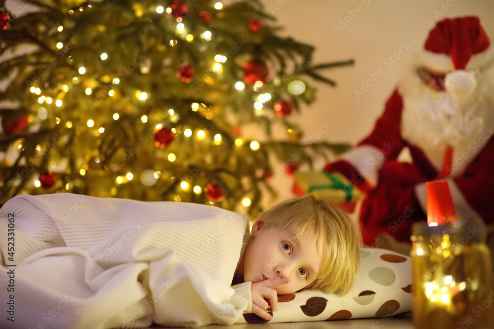 Little boy waiting Santa Claus under tree on Christmas Eve. Santa Claus brought gifts. Child not sleeping, but he does not show it. Magic at Christmas and New Year night. Little boy waiting Santa Claus under tree on Christmas Eve. Santa Claus brought gifts. Child not sleeping, but he does not show it. Magic at Christmas and New Year night.