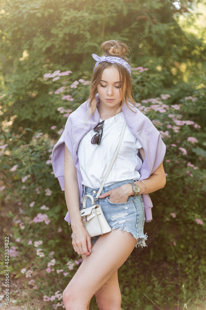 Beautiful hipster girl with a bandana in fashionable clothes with denim shorts, a purple sweatshirt and a white T-shirt stands near flowers Beautiful hipster girl with a bandana in fashionable clothes with denim shorts, a purple sweatshirt and a white T-shirt stands near flowers