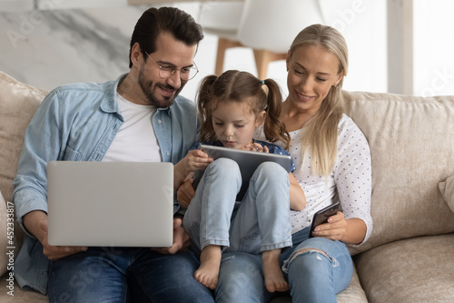 Happy family couple with little kid engaged in gadgets using, resting on sofa at home together. Parents watching daughter girl using app on tablet, playing online game, learning to read