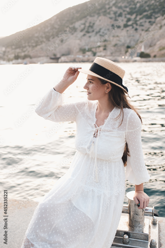 Stylish young woman 24-26 year old wear straw hat and white elegant dress posing over sea at quayside outdoors. Summer vacation season. Romantic lady relax at seashore.