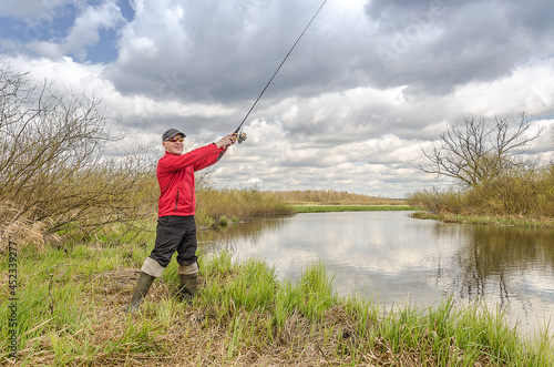 Fisherman in a red jacket is fishing on the green bank of the river.