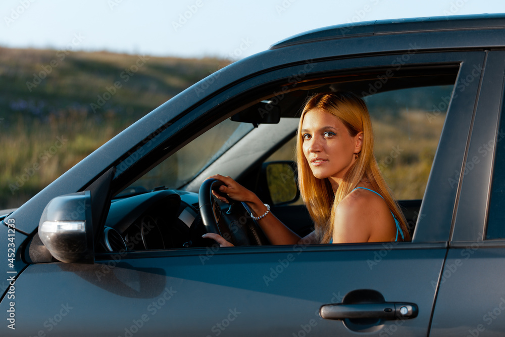 Naklejka premium Portrait of a young woman sitting behind the wheel of a car on a nature background with sunset light. Shallow focus.