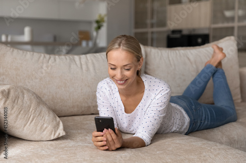 Happy woman using smartphone for online chat, enjoying virtual talk, making video call. User holding mobile phone, smiling, resting on sofa at home, watching media content on internet