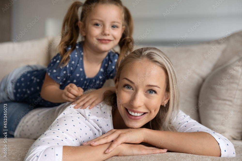 Portrait of happy beautiful mom with cute little daughter girl resting on her back. Mommy lying on couch at home, piggybacking child, enjoying leisure time, activity, playing games, looking at camera