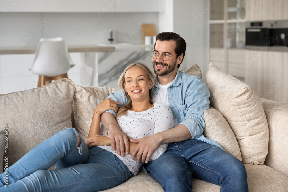 Happy dreamy millennial married couple resting and hugging on sofa at home, looking away, thinking over future family, house buying, mortgage, apartment rent. Smiling husband embracing wife
