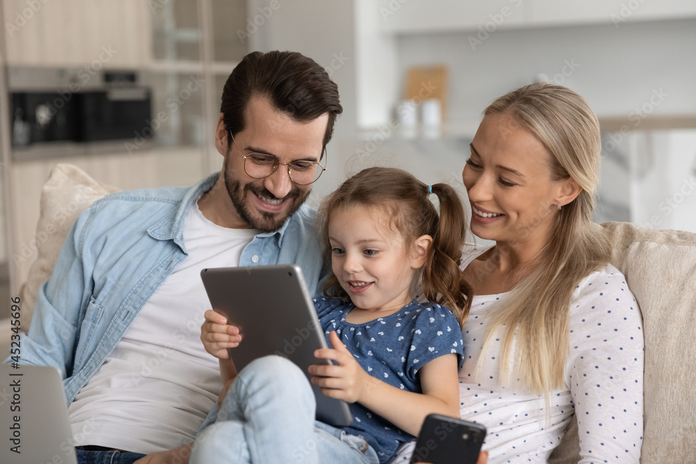 Happy excited gen Z little kid with tablet and parents with gadgets relaxing on couch, playing online game, looking at screen, smiling, laughing. Family using devices, making video call from home Happy excited gen Z little kid with tablet and parents with gadgets relaxing on couch, playing online game, looking at screen, smiling, laughing. Family using devices, making video call from home