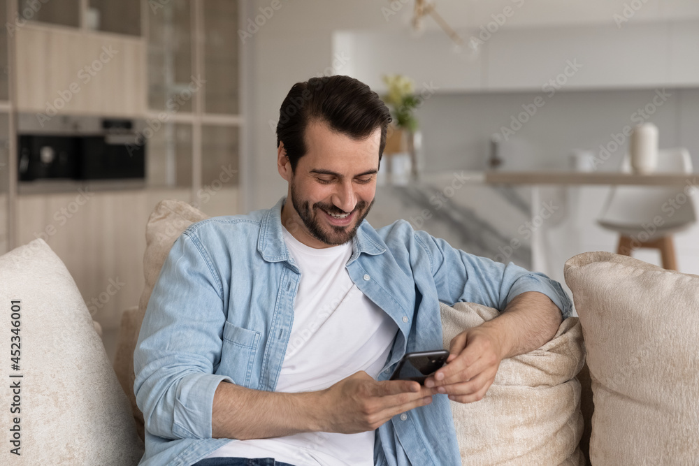 Happy cellphone user getting good news, reading text message on smartphone, smiling at screen. Millennial man using mobile phone on couch at home, shopping online, chatting on social media