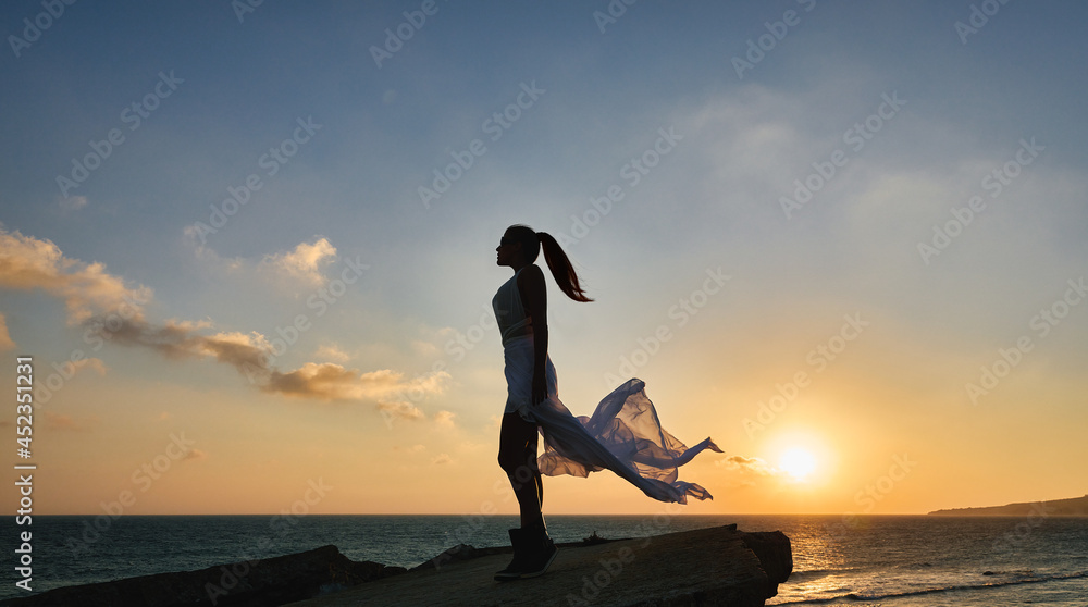 Trendy and sexy beautiful woman in a white swimsuit and flowing sarong silhouetted against the colorful orange tropical sunset reflected on the ocean