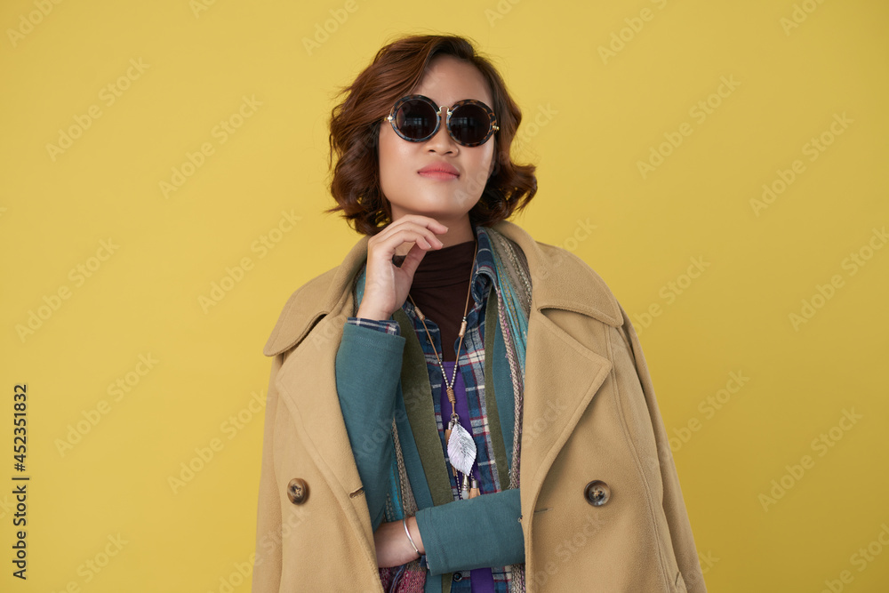 Portrait of stylish fashionable young woman in warm coat and sunglasses standing against yellow background