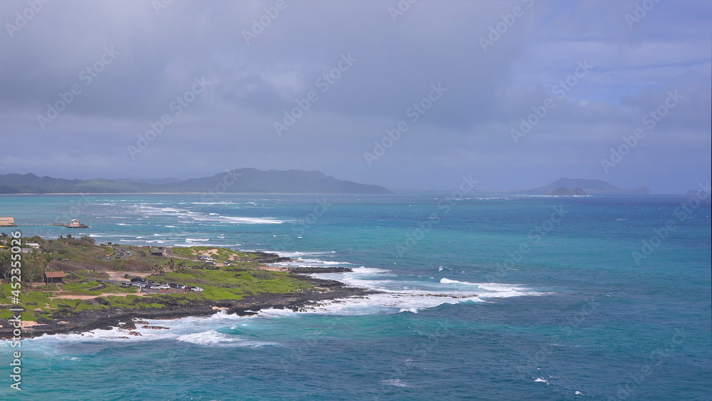 View of Makapuu beach. Waves of Pacific Ocean wash over yellow sand of tropical beach. Magnificent mountains of Hawaiian island of Oahu against backdrop of blue sky with white clouds. View of Makapuu beach. Waves of Pacific Ocean wash over yellow sand of tropical beach. Magnificent mountains of Hawaiian island of Oahu against backdrop of blue sky with white clouds.