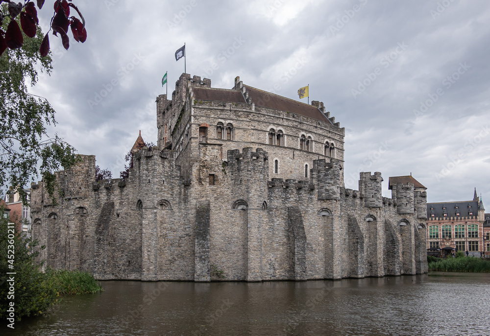 Gent, Flanders, Belgium - July 30, 2021: Gray stone Gravensteen medieval castle under gray cloudscape behind its dark water moat. Some green foliage and colored flags.