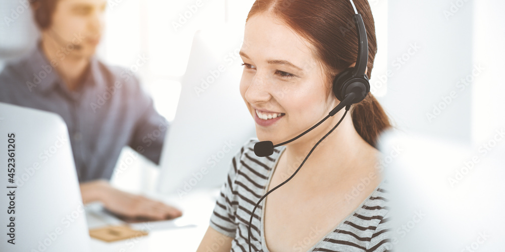 Casual dressed young woman using headset and computer while talking with customers online. Group of operators at work in sunny office. Call center Casual dressed young woman using headset and computer while talking with customers online. Group of operators at work in sunny office. Call center