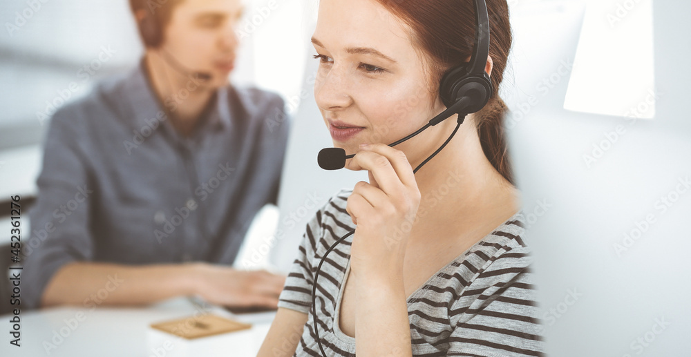 Casual dressed young woman using headset and computer while talking with customers online. Group of operators at work in sunny office. Call center Casual dressed young woman using headset and computer while talking with customers online. Group of operators at work in sunny office. Call center