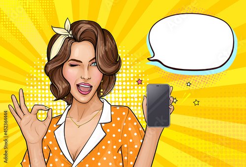 Pop art girl holding mobile phone. Winking woman showing screen of new smartphone and OK sign. Pretty lady on yellow background with empty speech bubble. Vector illustration for digital advertisement.