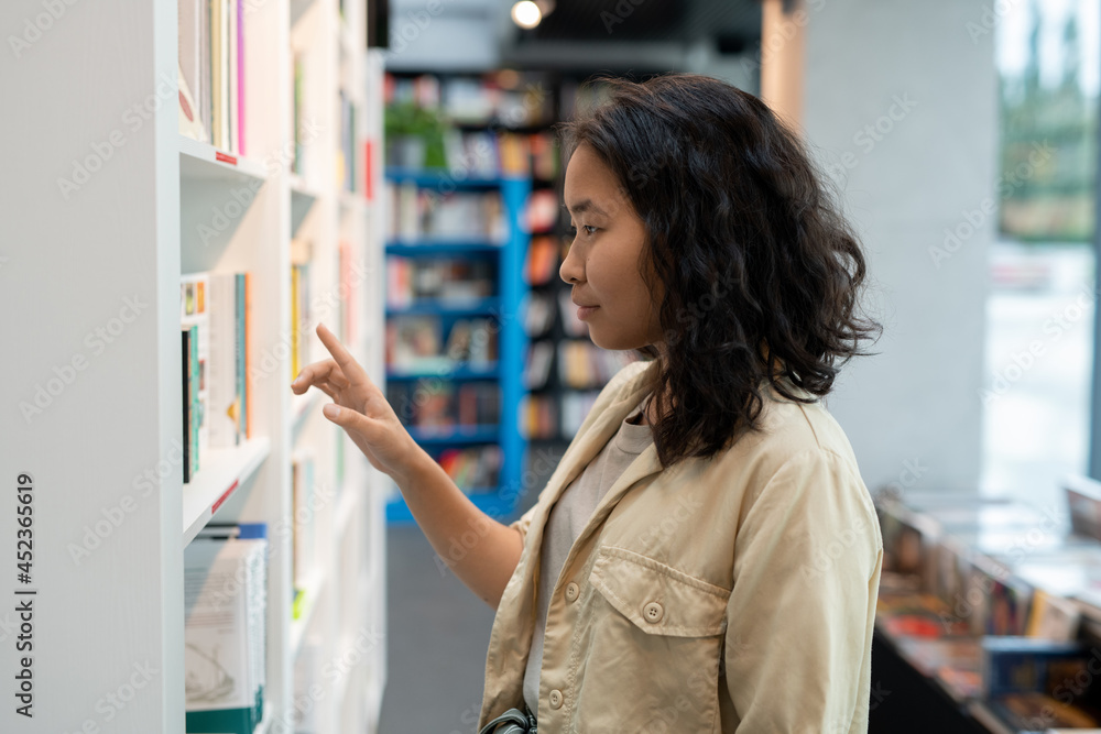 Fototapeta premium Young businesswoman or student standing by one of shelves with books in library