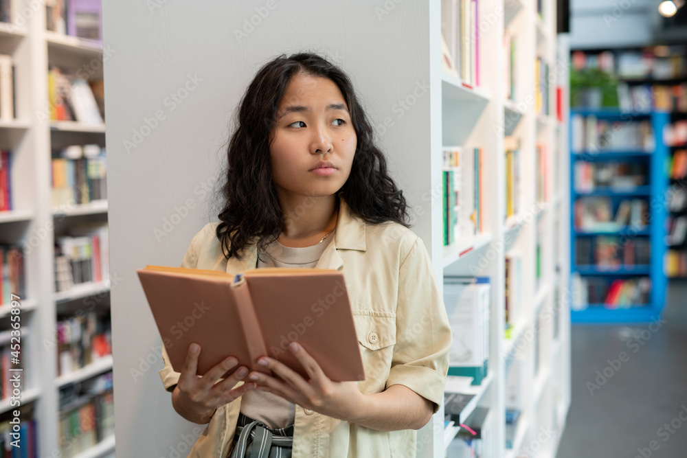 Young Asian woman holding open book while standing against bookshelf in library Young Asian woman holding open book while standing against bookshelf in library