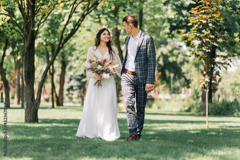 Happy young groom and beautiful tender bride in a wedding dress with a bouquet, Bride and groom hold hands and walk on a green field Happy young groom and beautiful tender bride in a wedding dress with a bouquet, Bride and groom hold hands and walk on a green field