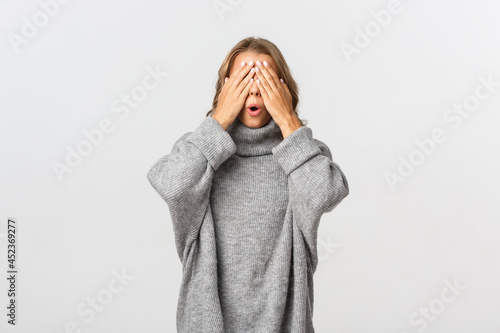 Portrait of beautiful young woman in grey sweater, standing blindsided with hands on face, waiting for surprise, standing over white background photo