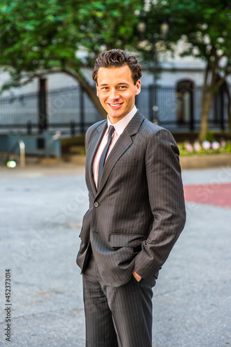 Wearing a black striped jacket, tie, two hands putting in pockets,  a young businessman is standing outside,  smiling,  looking at you.. © Alexander Image