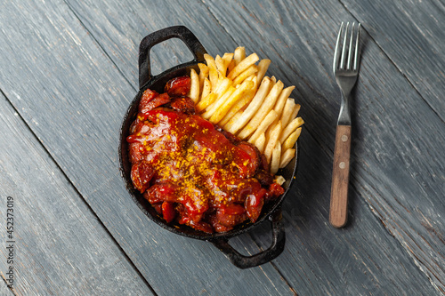 Currywurst with french fries in black pan on wooden table. Top view.